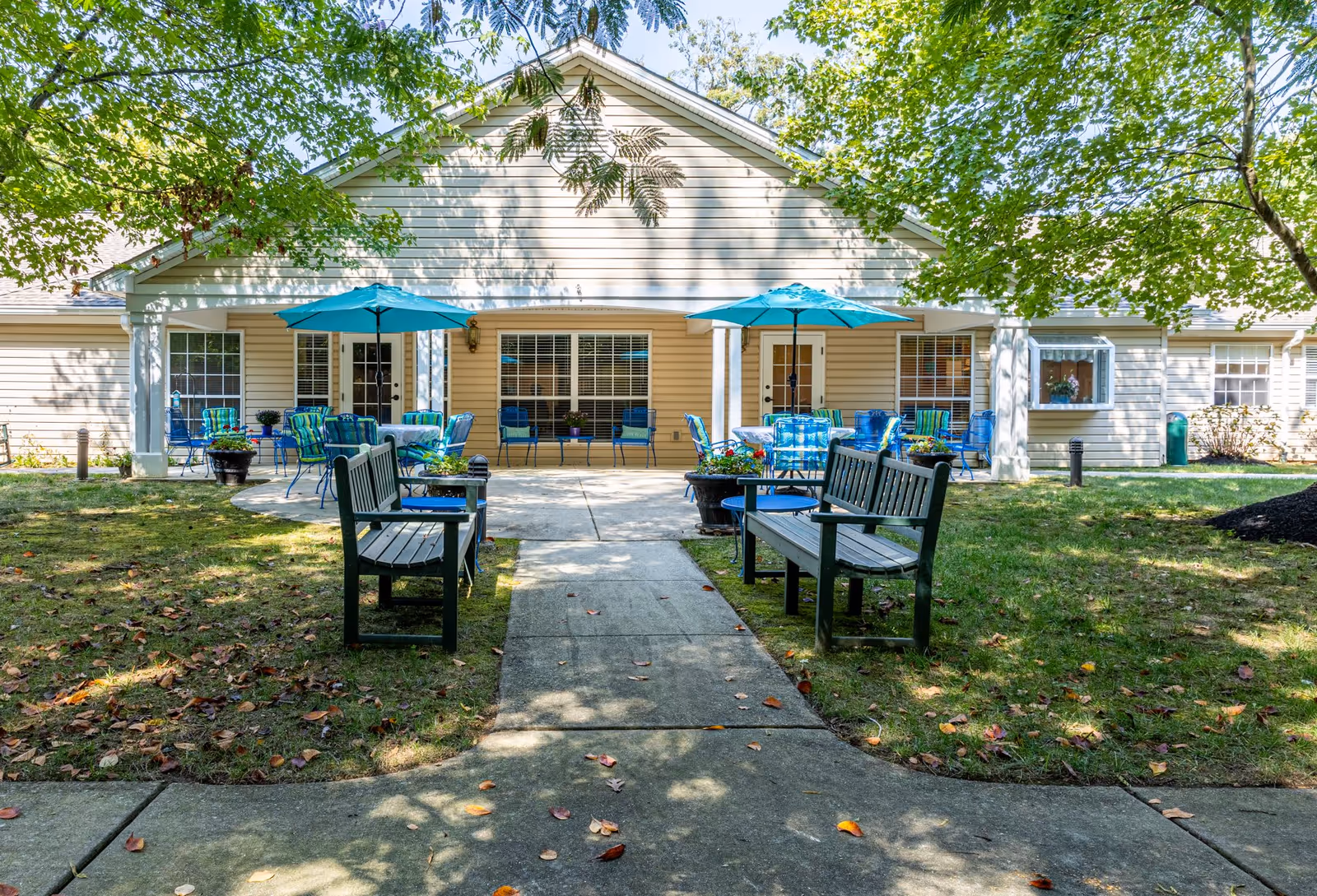 Outdoor patio area at Charter Senior Living of Annapolis featuring a concrete walkway flanked by wooden benches leading to a building with beige siding. The patio has multiple tables with blue umbrellas and chairs, surrounded by green grass and trees providing shade.