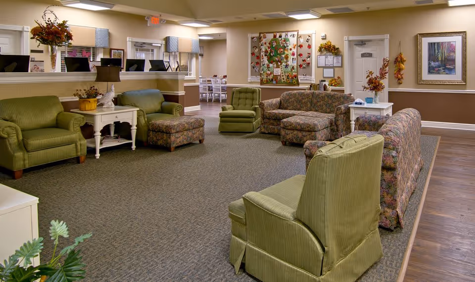 A spacious senior living facility common area with multiple upholstered armchairs and sofas in green and floral patterns arranged around a carpeted floor. There are side tables with decorative items and flowers, a bulletin board with colorful decorations on the wall, and a doorway leading to a dining area with tables and chairs. The walls are painted beige with a brown lower section, and there is a framed painting on the wall.