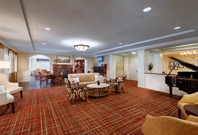 Spacious senior living facility common area with patterned red carpet, beige sofa, two patterned armchairs, a round coffee table, a grand piano, and a dining area with tables and chairs near a fireplace in the background.