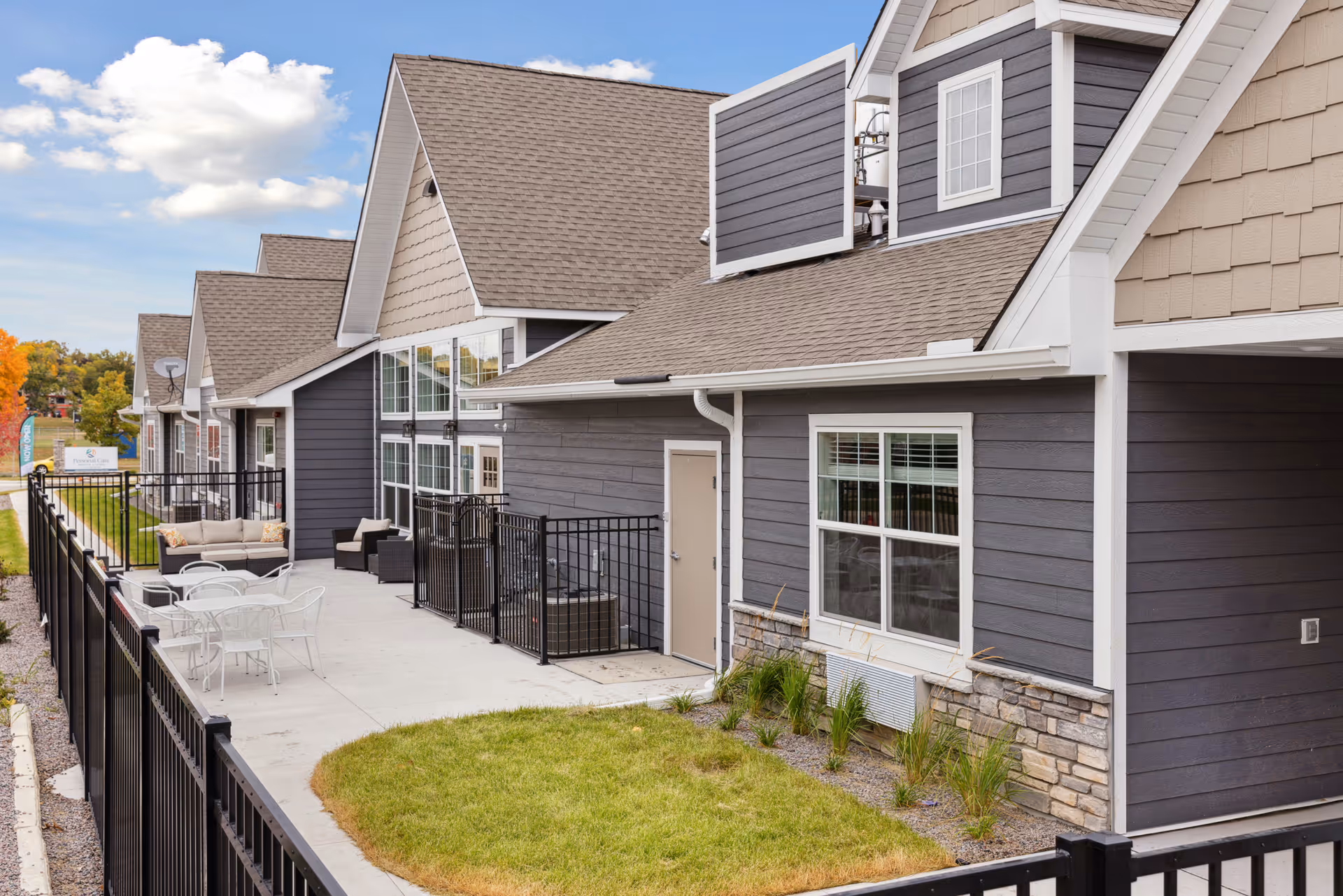Exterior view of a senior living facility building with gray siding, multiple windows, and a sloped roof. There is a fenced patio area with outdoor seating including cushioned sofas and white metal tables and chairs. The sky is partly cloudy and there is some greenery around the building.