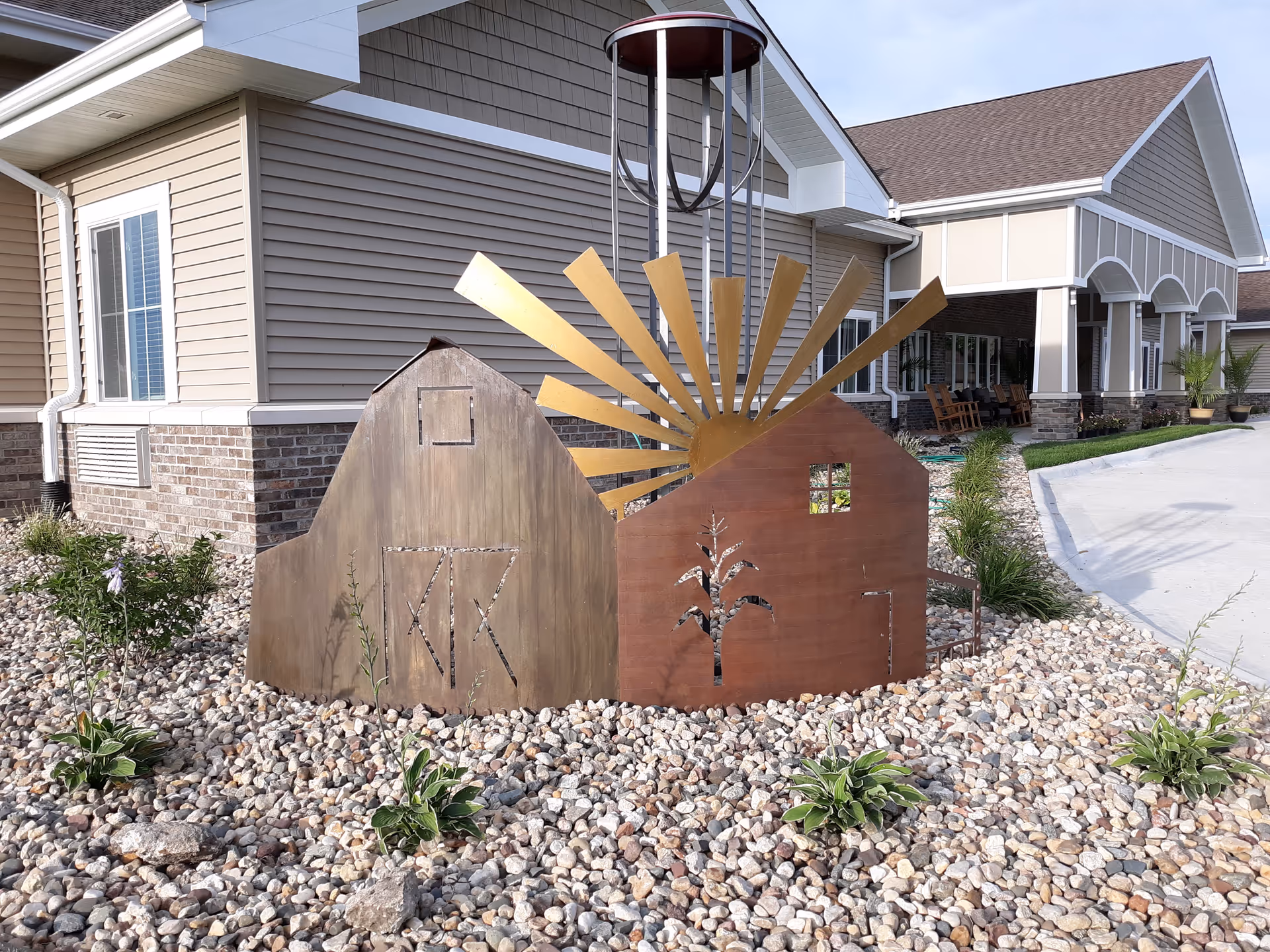 Outdoor view of a senior living facility named Country Meadow Place featuring a decorative metal sculpture of two barns with a sun rising behind them, surrounded by small plants and rocks, with the building and entrance visible in the background.