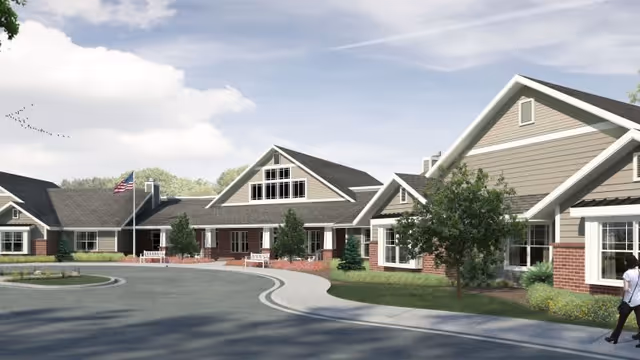 Exterior view of a single-story senior living facility building with beige siding and brick accents, multiple peaked roofs, an American flag on a flagpole, landscaped greenery, and a curved driveway. A person is walking on the sidewalk near the building.