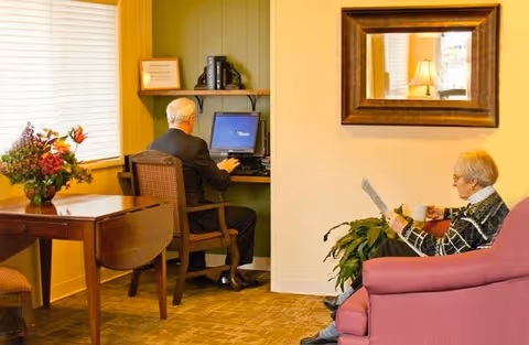 Two elderly residents in a cozy common room, one using a desktop computer at a small desk and the other seated on a sofa reading with a mug.