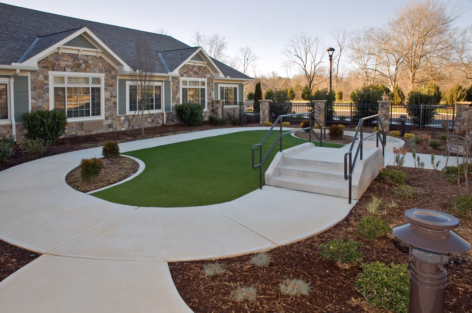 Outdoor area of a senior living facility featuring a stone and siding building with multiple windows, a curved concrete walkway surrounding a small patch of artificial grass, landscaped garden beds with shrubs, and a small set of concrete stairs with metal handrails leading to a fenced area.