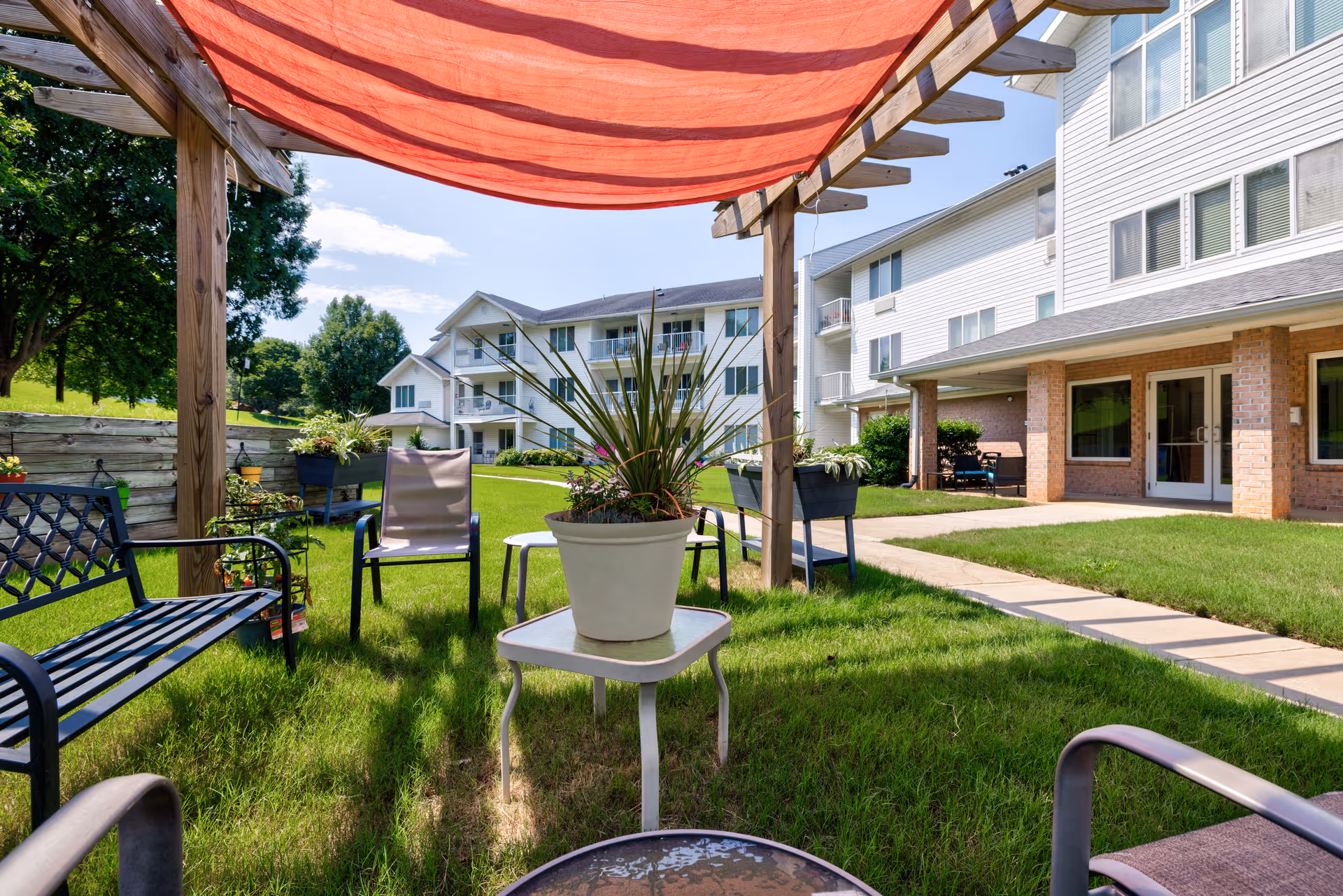 Outdoor seating area at TerraBella Epps Bridge with metal chairs and benches arranged on green grass under a wooden pergola with a red fabric canopy. In the background, there are multi-story residential buildings with balconies and windows, and a paved walkway runs through the grassy area.
