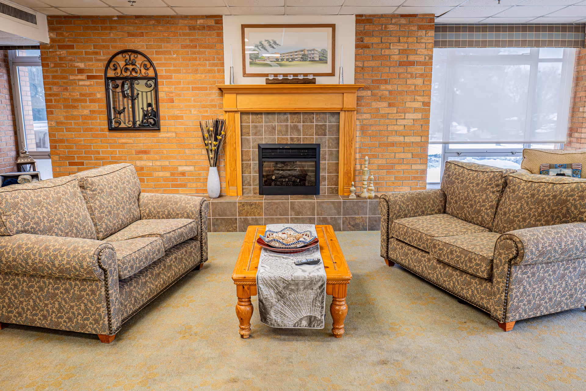 A cozy living room with two patterned sofas facing each other and a wooden coffee table with a decorative runner and a remote control on it. The room features a brick wall with a fireplace in the center, a framed picture above the mantel, and a decorative wall mirror on the left side. Large windows with blinds allow natural light to enter the space.
