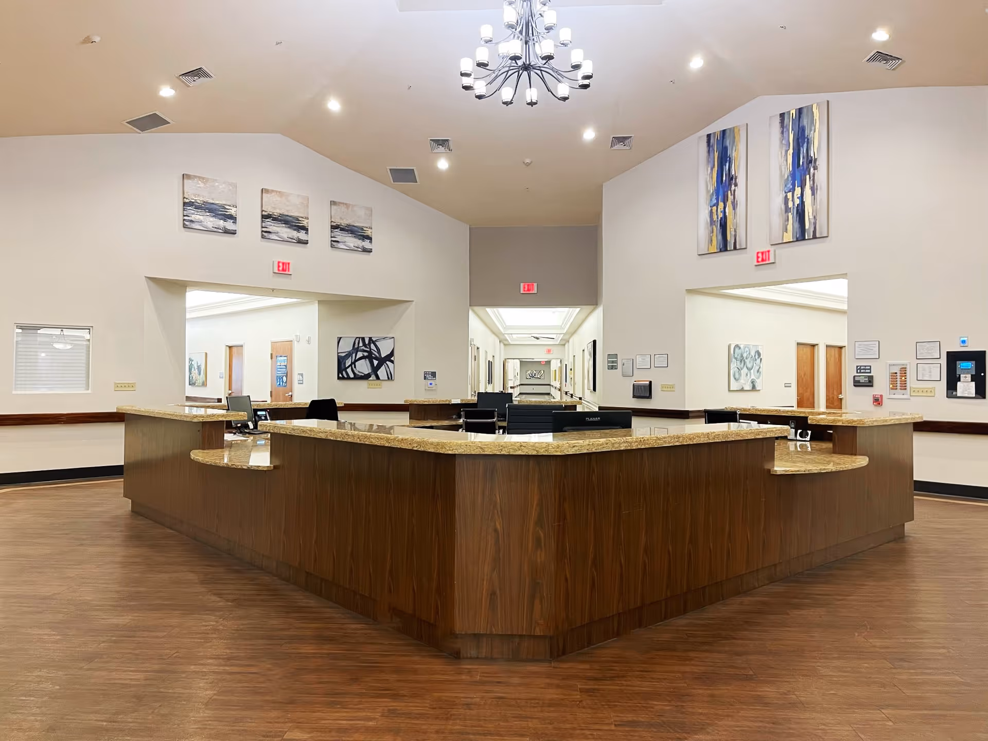 A spacious reception or nurse station area inside La Hacienda de Paz Rehabilitation and Care Center with a large wooden desk with granite countertops in the center. The room has high ceilings with a chandelier, light-colored walls adorned with abstract paintings, and multiple exit signs above doorways leading to hallways.