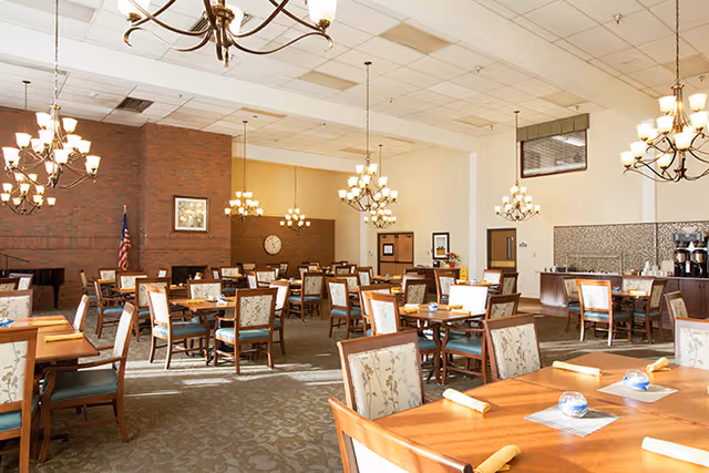 A spacious dining room with multiple wooden tables and chairs arranged neatly. The chairs have floral upholstery. The room features several chandeliers hanging from the ceiling, a brick wall with an American flag and framed artwork, and a clock on the far wall. There is a piano in the corner and a counter area with coffee machines on the right side.
