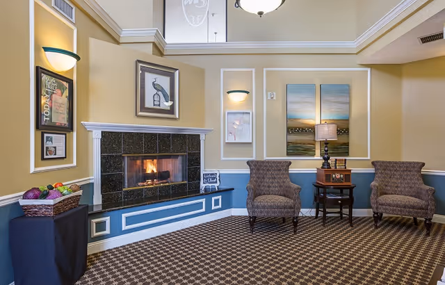 A cozy seating area in a memory care facility featuring a lit fireplace with a black tile surround, two patterned armchairs, a small wooden side table with a lamp and books, and framed artwork on the walls. A basket of artificial fruits is placed on a covered table to the left.