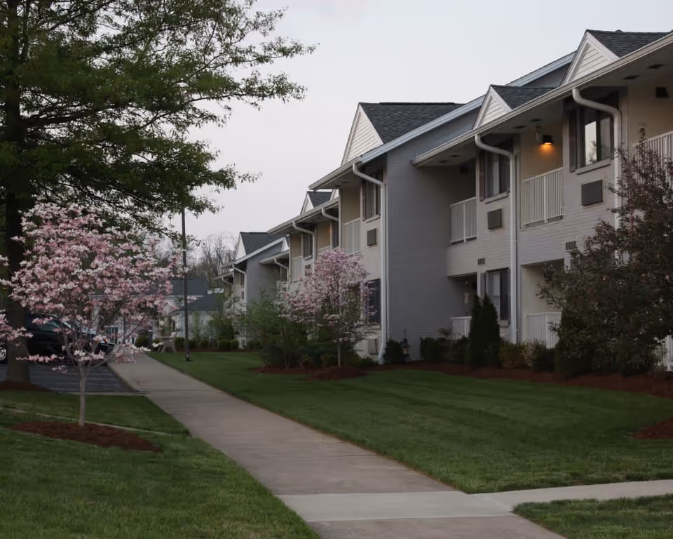 Exterior view of a senior living facility with a sidewalk running alongside a row of two-story buildings. The buildings have balconies and are surrounded by well-maintained green lawns and blooming pink trees. The sky is overcast.