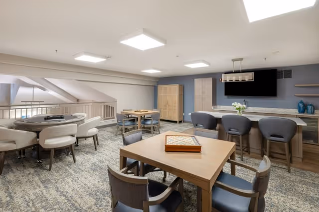 A spacious, well-lit common area with multiple tables and chairs arranged for games and socializing. The room features a large flat-screen TV mounted on the wall above a countertop with bar stools. The decor includes neutral tones with blue accents and modern lighting fixtures on the ceiling.