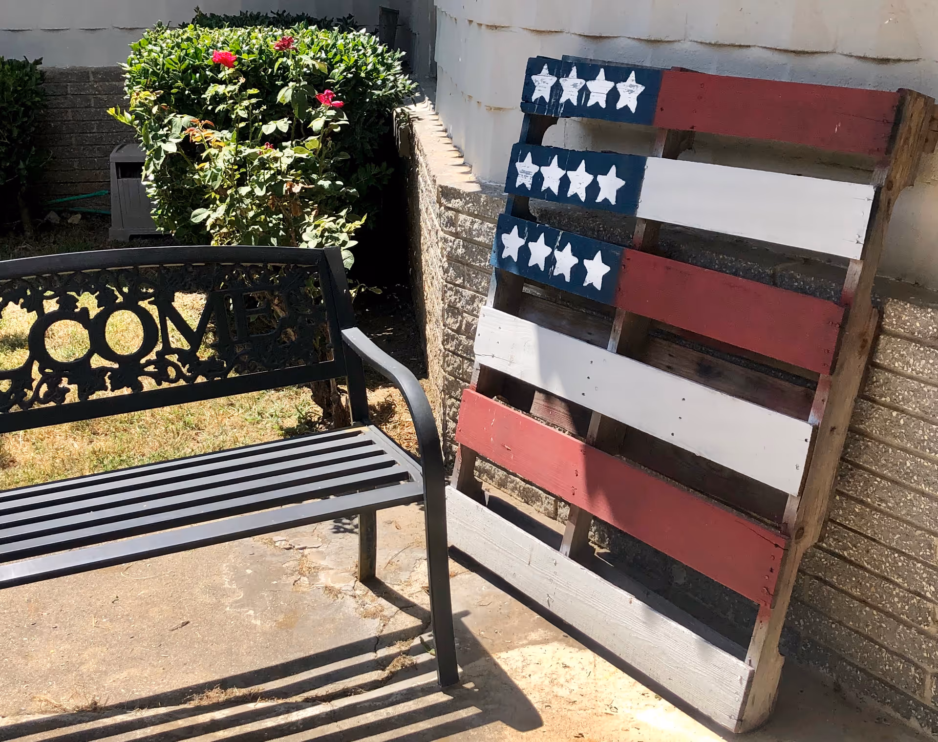 A metal welcome bench sits beside a wooden pallet painted like an American flag leaning against an exterior wall near shrubs.