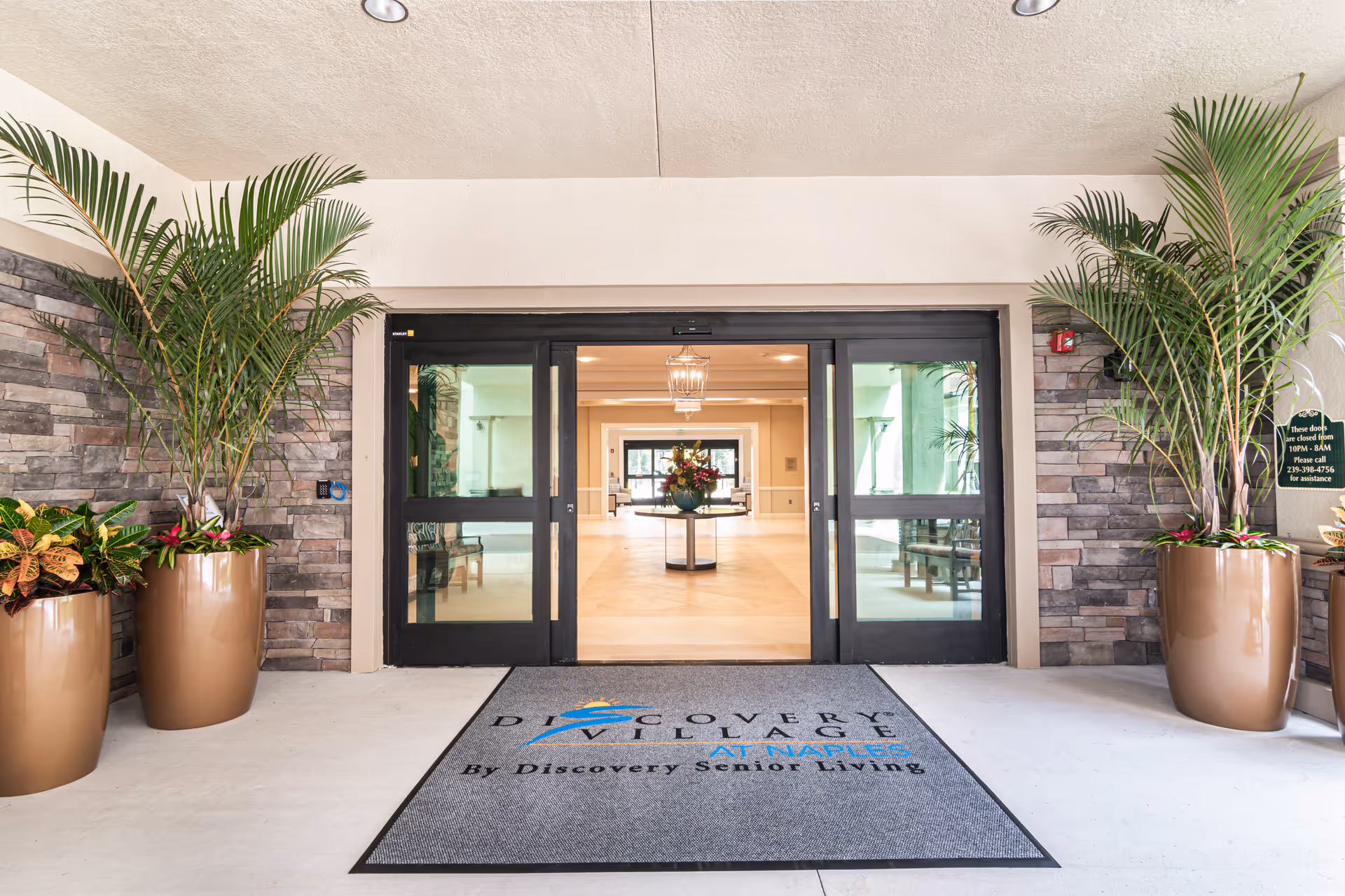 Entrance to Discovery Village At Naples senior living facility with automatic sliding glass doors, large potted plants on either side, and a welcome mat with the facility's name. Inside, a round table with a floral arrangement is visible in a well-lit lobby area.