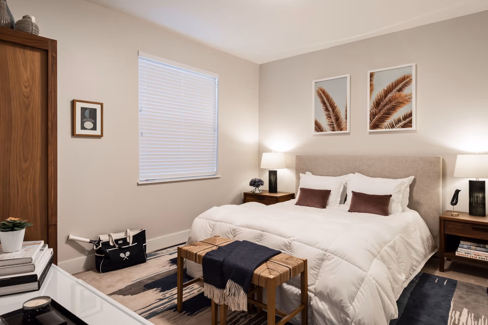 A cozy bedroom featuring a large bed with white bedding and two purple accent pillows. On either side of the bed are wooden nightstands with lamps and decorative items. Above the bed are two framed pictures of palm leaves. A woven bench with a dark throw blanket is placed at the foot of the bed. The room has a window with white blinds, a wooden wardrobe, and a patterned rug on the floor.