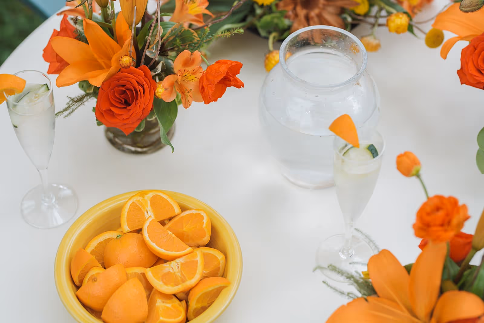 A white table with a yellow bowl filled with sliced oranges, a clear glass pitcher of water, two glasses with water garnished with cucumber slices and orange peels, and orange floral arrangements including roses and lilies.