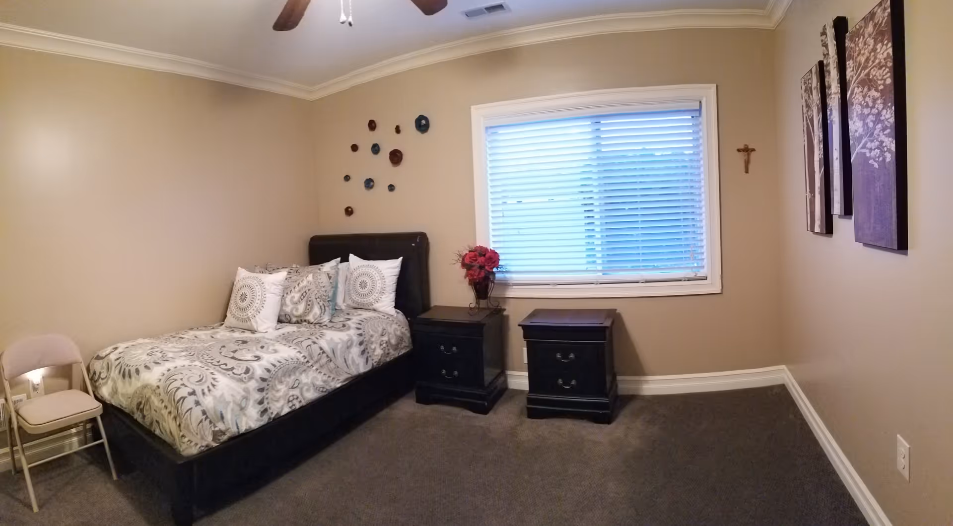 A bedroom with a bed covered in patterned bedding and three decorative pillows. There are two black nightstands next to the bed, one with a vase of red flowers. A window with white blinds is on the wall behind the nightstands. The walls are beige with decorative wall art and a small cross. A beige folding chair is placed near the bed. The room has carpeted flooring and a ceiling fan with lights.