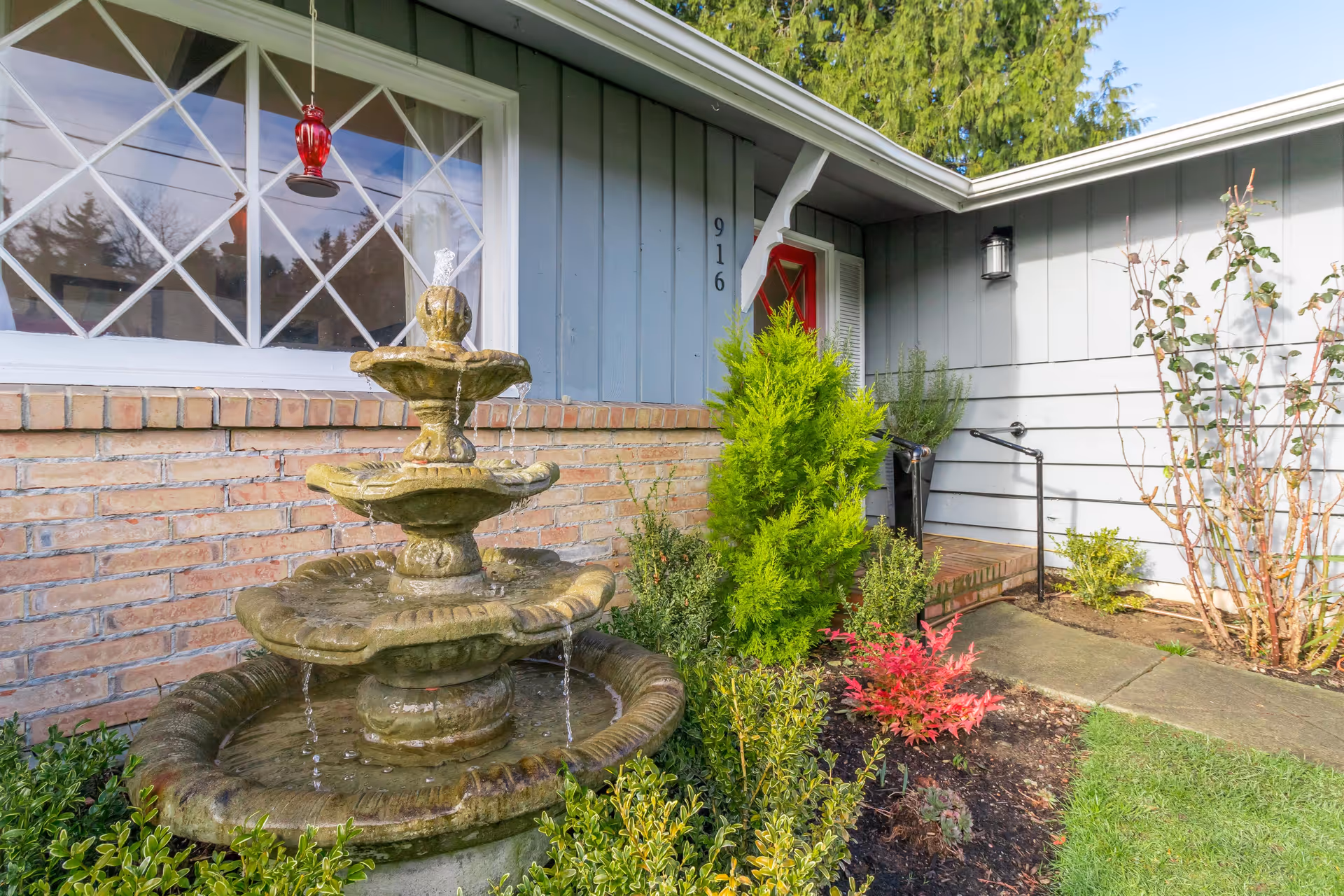 Front exterior of a house with a three-tier stone fountain in the landscaped yard and a red entry door under a small porch.