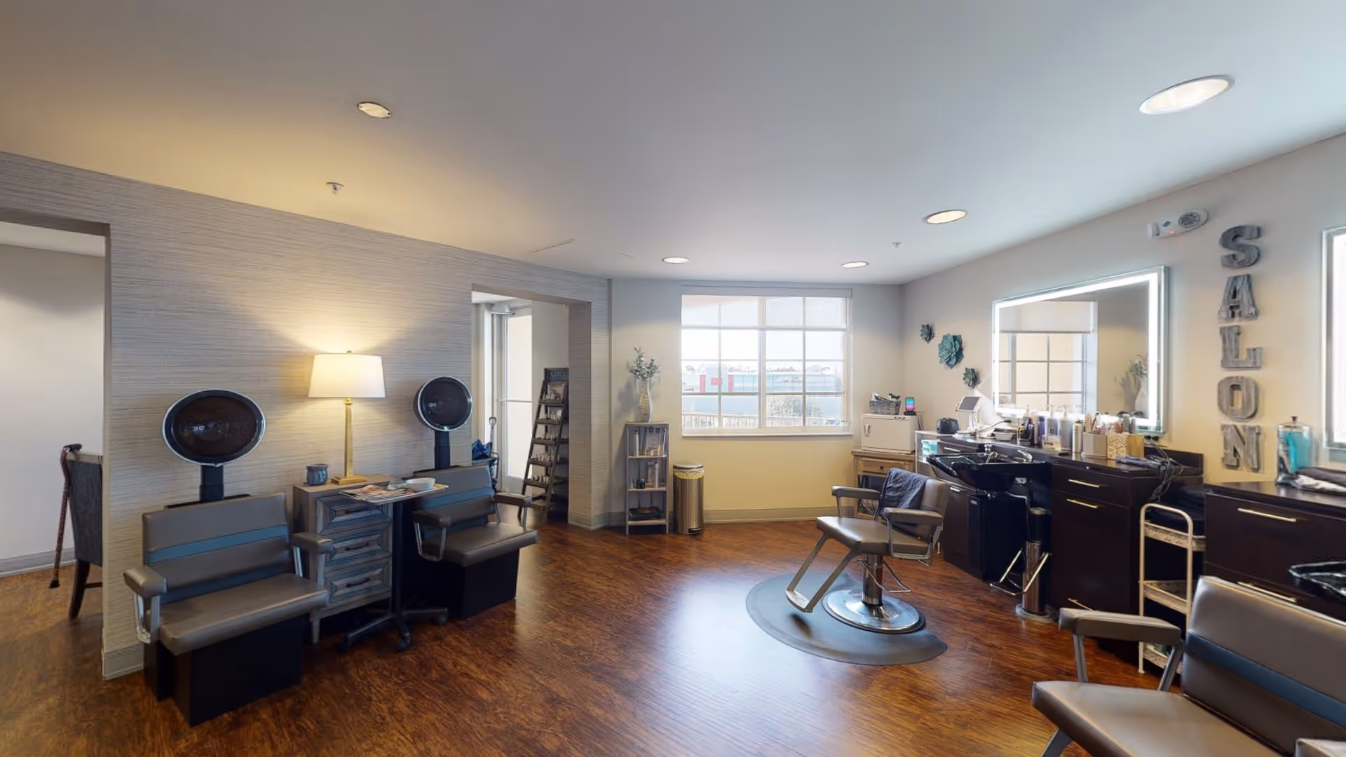 Interior view of a salon room in a senior living facility with salon chairs, hair dryers, a large mirror with lights, and various hair care products on counters. The room has wooden flooring, a window letting in natural light, and decorative wall art spelling SALON.