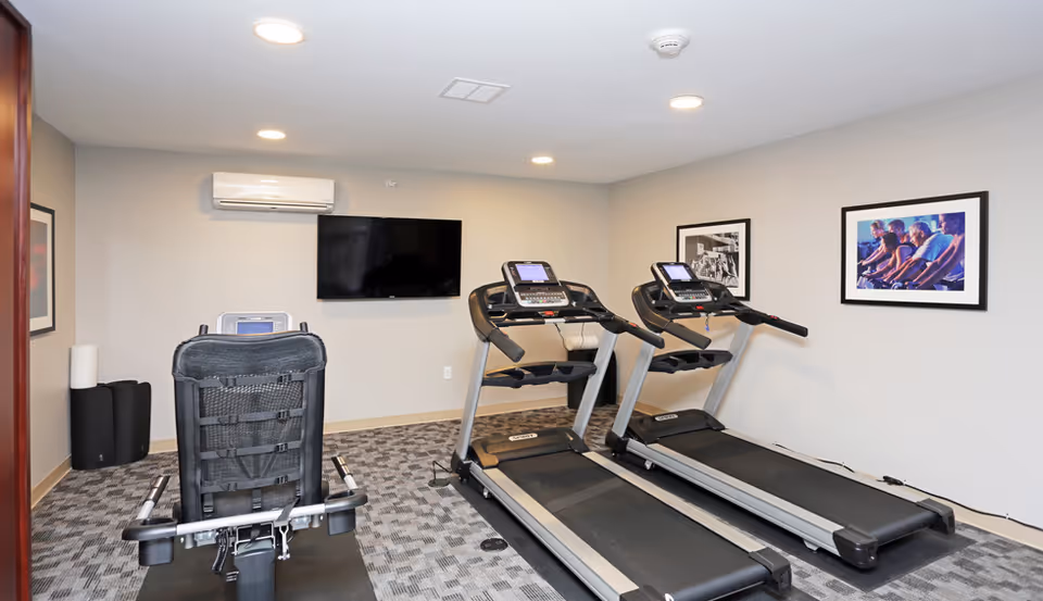 Small fitness room with two treadmills, a recumbent bike, a wall-mounted TV, and framed pictures on the walls.