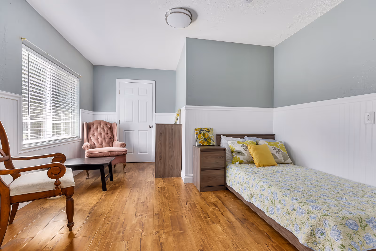 Well-lit bedroom with a single bed and floral bedspread, a nightstand, wooden chairs, and a pink upholstered armchair by a window.