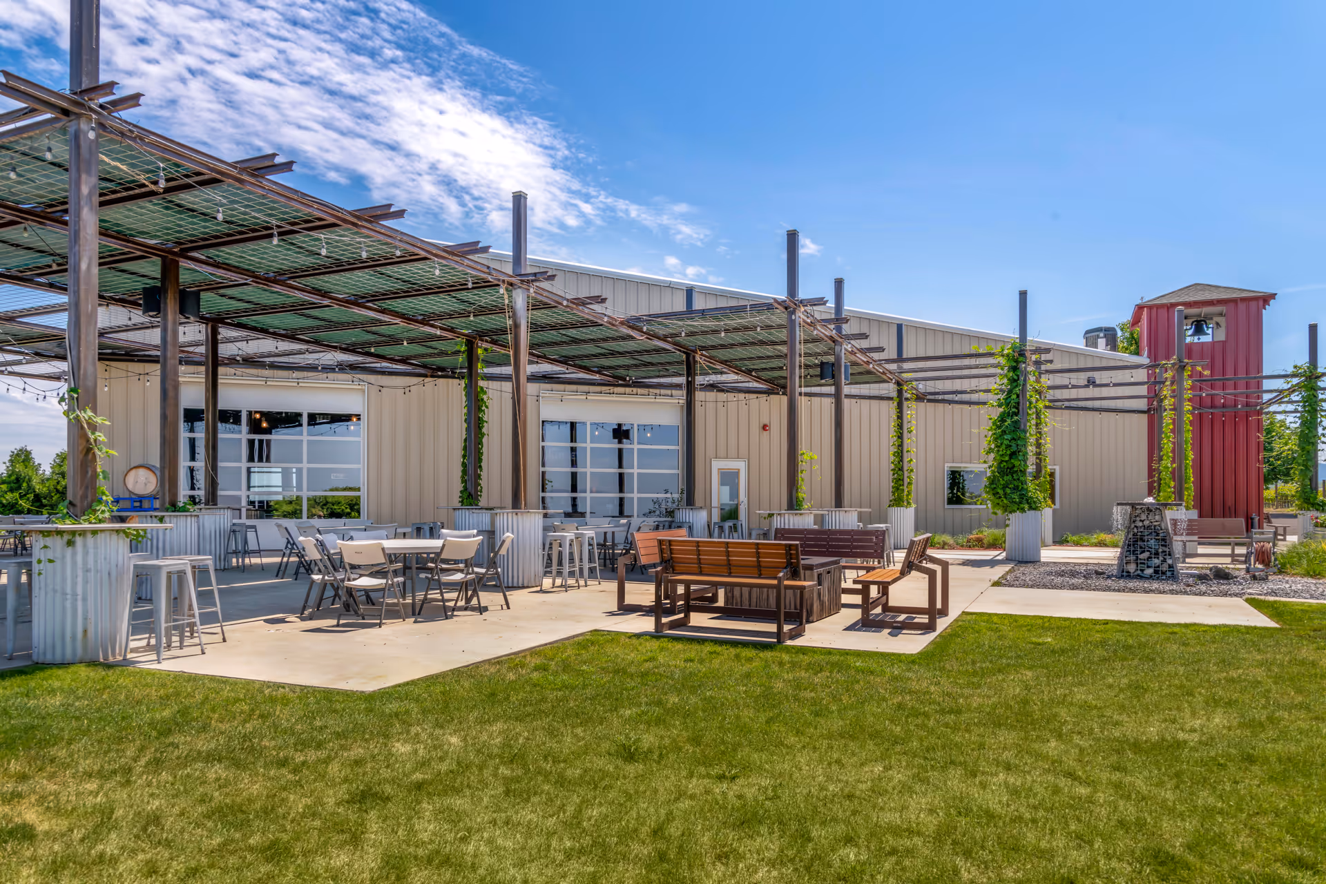 Outdoor patio area with metal pergolas covered in greenery, several tables and chairs, wooden benches, and a fire pit, adjacent to a large beige building under a blue sky with some clouds.