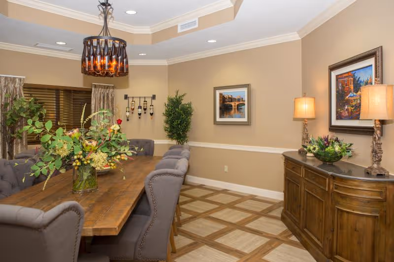 A dining room with a long wooden table surrounded by upholstered chairs. The table has a large floral arrangement in a glass vase. The room features beige walls with white trim, two framed paintings, a wooden sideboard with two lamps and a decorative bowl, a wine rack on the wall, and a potted plant in the corner. A chandelier with multiple lights hangs above the table.