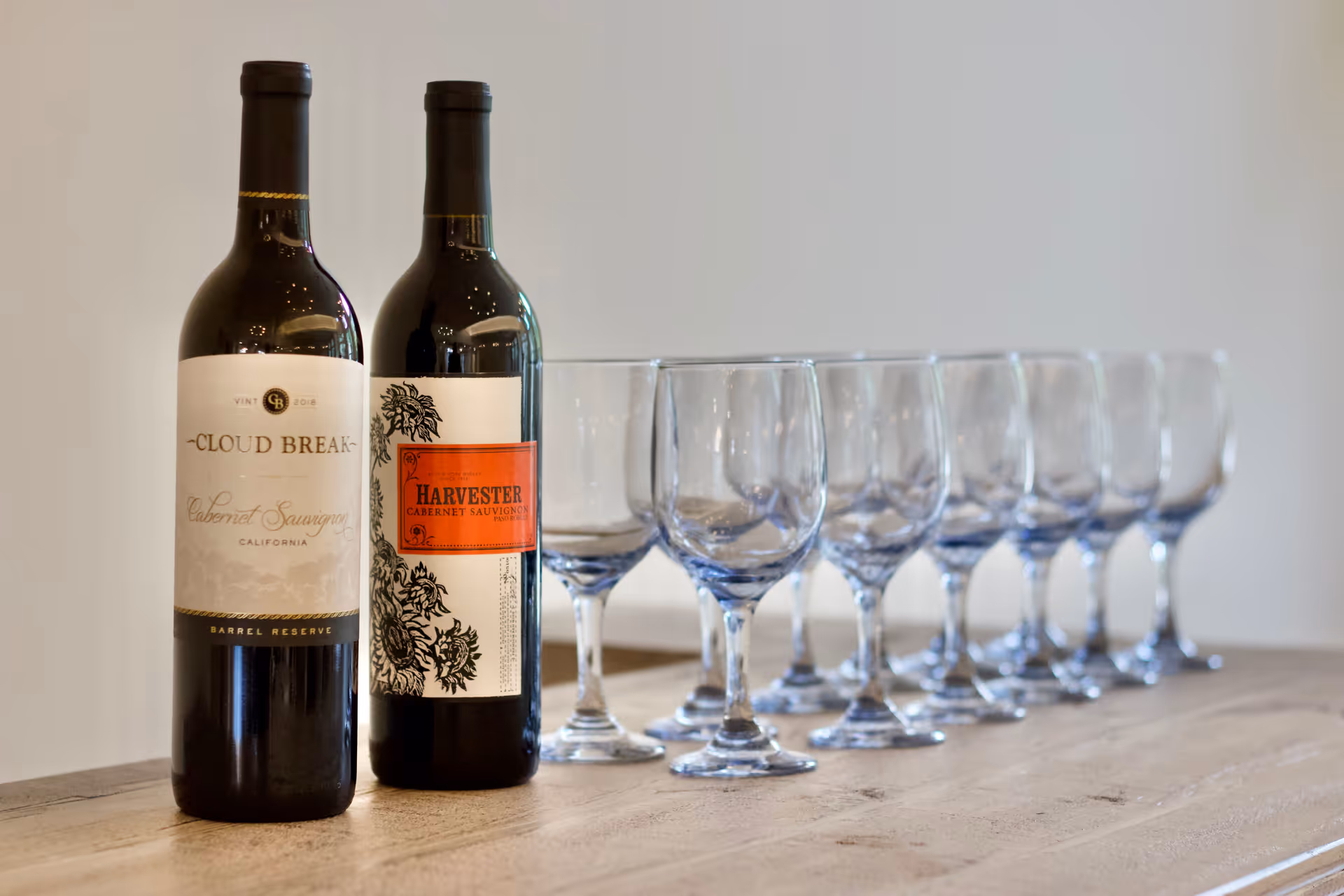 Two bottles of red wine, one labeled Cloud Break Cabernet Sauvignon and the other labeled Harvester Cabernet Sauvignon, placed on a wooden surface next to a row of eight empty wine glasses.