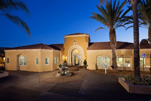 Exterior view of a single-story building at dusk with a central entrance featuring an arched doorway, a small fountain in front, palm trees, and well-lit windows. The building has a beige stucco finish and a tiled roof.