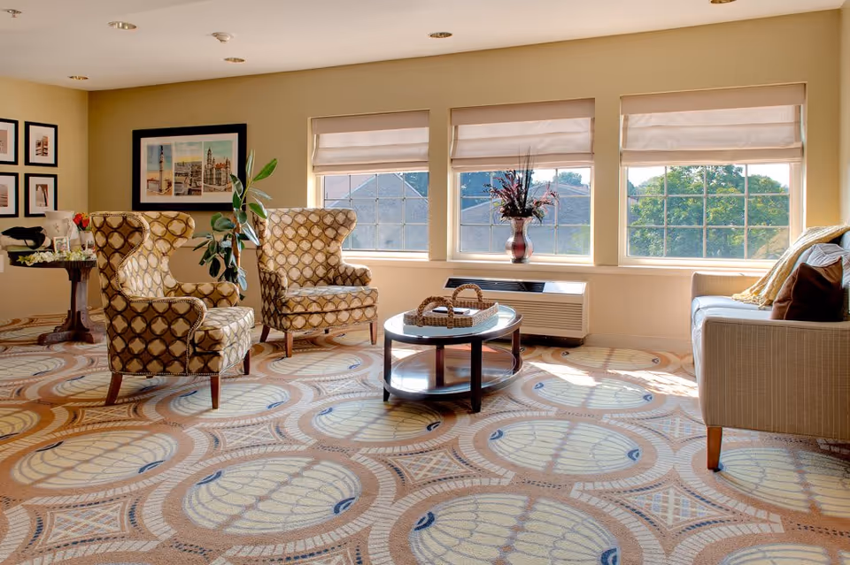 A bright and cozy living room with patterned carpet, two upholstered armchairs with a circular pattern, a beige sofa with pillows, a round wooden coffee table with a decorative basket, and three large windows with beige blinds letting in natural light. There are framed pictures on the wall and a vase with flowers on the windowsill.