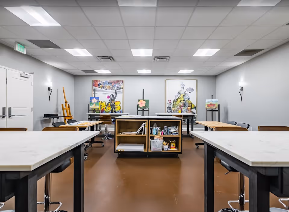 Well-lit art studio with worktables, easels, paintings, and supply shelves.