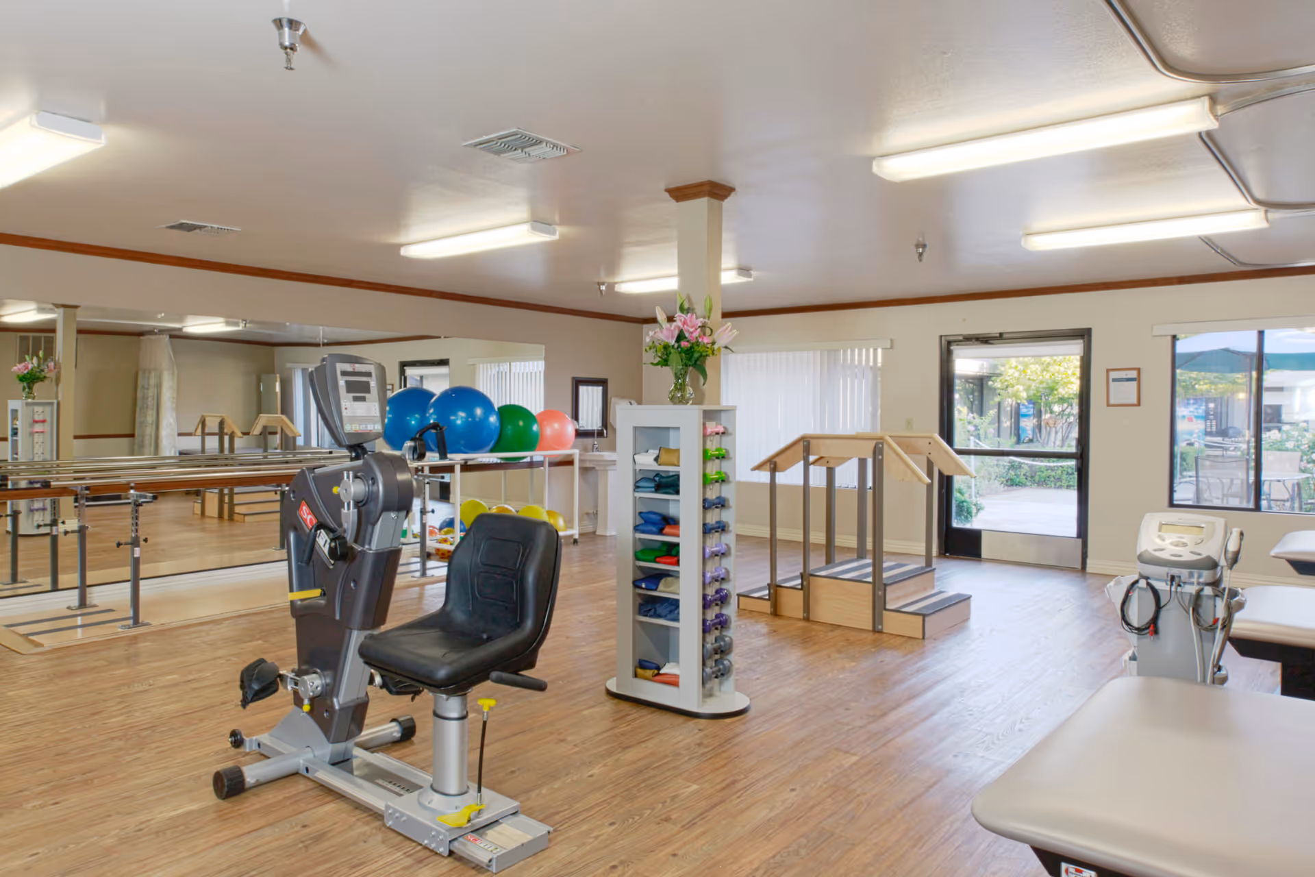 A physical therapy room with exercise equipment including a recumbent bike, colorful exercise balls, hand weights on a rack, parallel bars, and a small set of stairs with handrails. The room has wooden flooring, large mirrors on one wall, and windows showing an outdoor patio area.