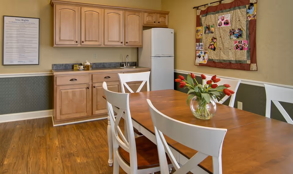 Dining area with a wooden table and white chairs, a vase of red tulips, and a small kitchenette with cabinets and a refrigerator.