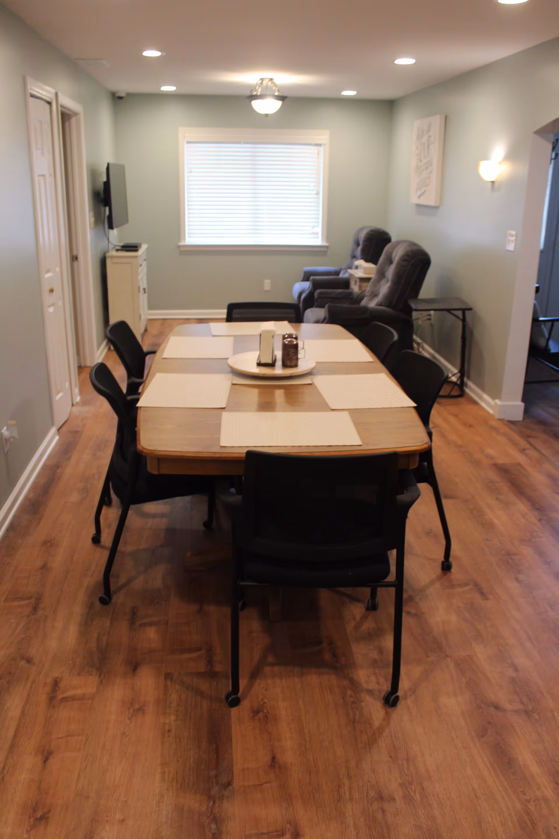 A dining area with a wooden table set with six black chairs, each with a beige placemat. On the table is a round tray holding a napkin holder and a small container. In the background, there are two gray recliner chairs next to a small side table, a window with closed blinds, and a wall-mounted TV on the left. The room has wooden flooring and light blue walls with ceiling and wall lights.