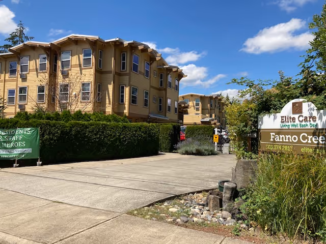 Exterior view of a multi-story senior living facility building with beige siding and multiple windows under a blue sky with some clouds. There is a concrete driveway and landscaped greenery including bushes and trees. A sign in the foreground reads 'Elite Care Living Well Each Day Fanno Creek' with contact information.
