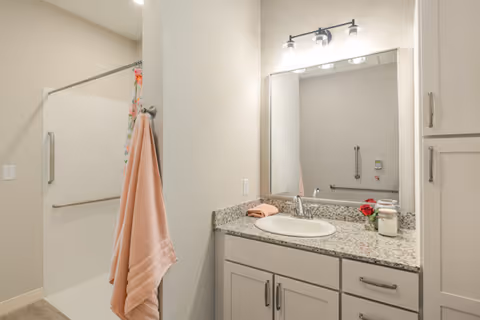 A clean and modern bathroom featuring a granite countertop with a sink, a large mirror with three light fixtures above it, a pink towel hanging on a hook, and a walk-in shower with a glass door and grab bars.