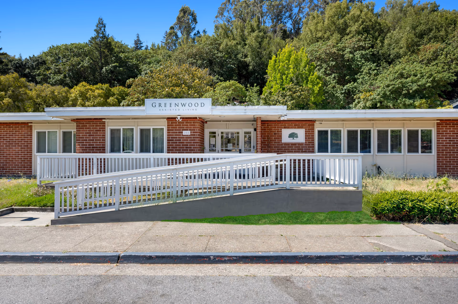 Front exterior of Greenwood Assisted Living, a single-story brick building with a white wheelchair ramp and trees behind it.