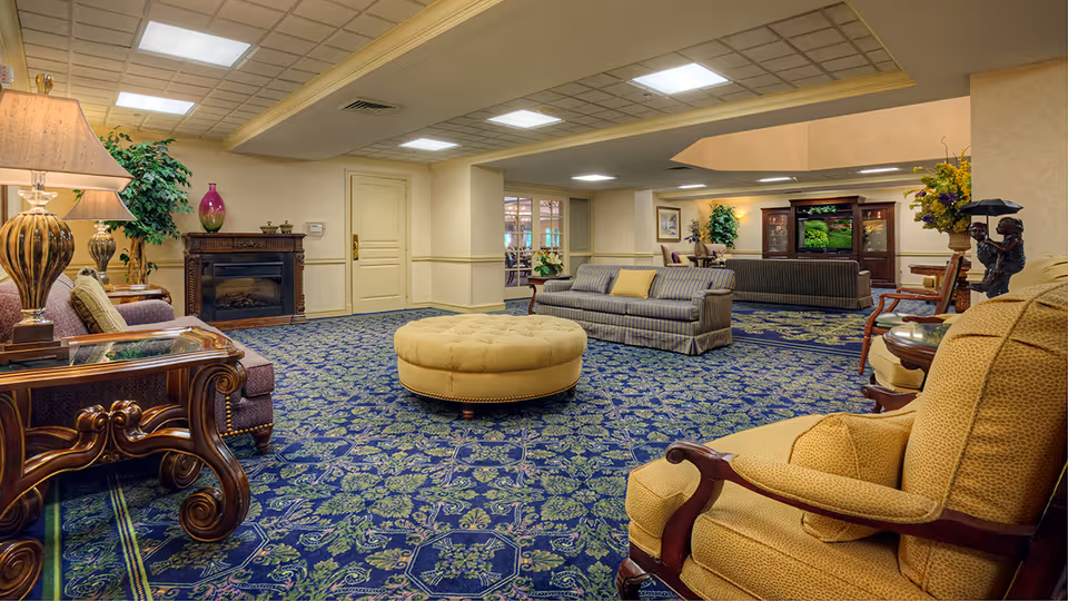 A spacious senior community lounge area with patterned blue and green carpet, multiple upholstered sofas and armchairs, a round yellow ottoman in the center, decorative lamps, plants, and a large wooden entertainment center with a television.
