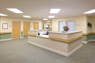 Interior view of a senior living facility reception area with a central desk, beige walls, green lower wall paneling, multiple closed wooden doors, overhead fluorescent lighting, and a vase with flowers on the desk.