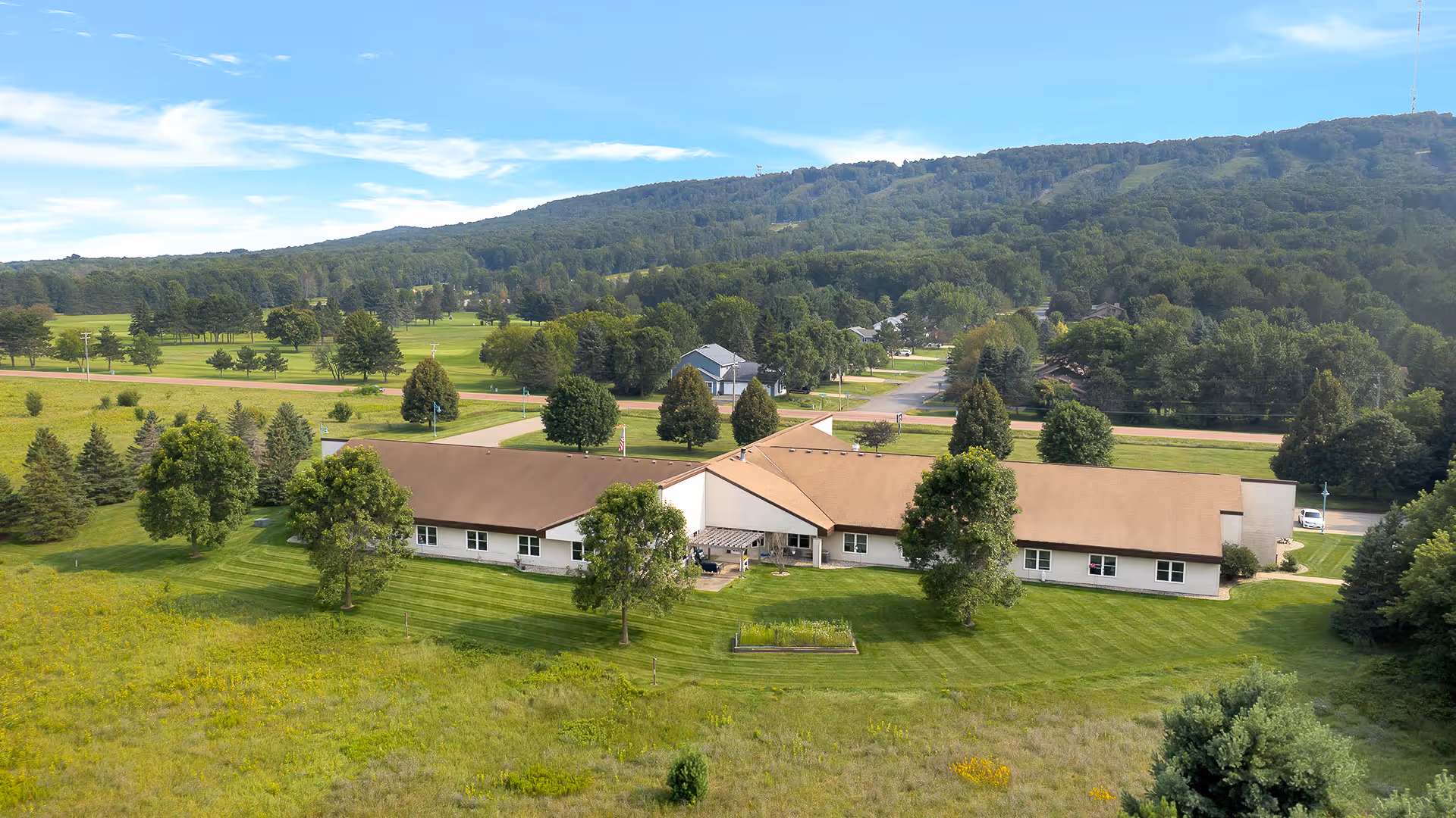 Aerial view of a single-story senior living building with a brown roof surrounded by mowed lawns, trees, and rolling hills in the background.