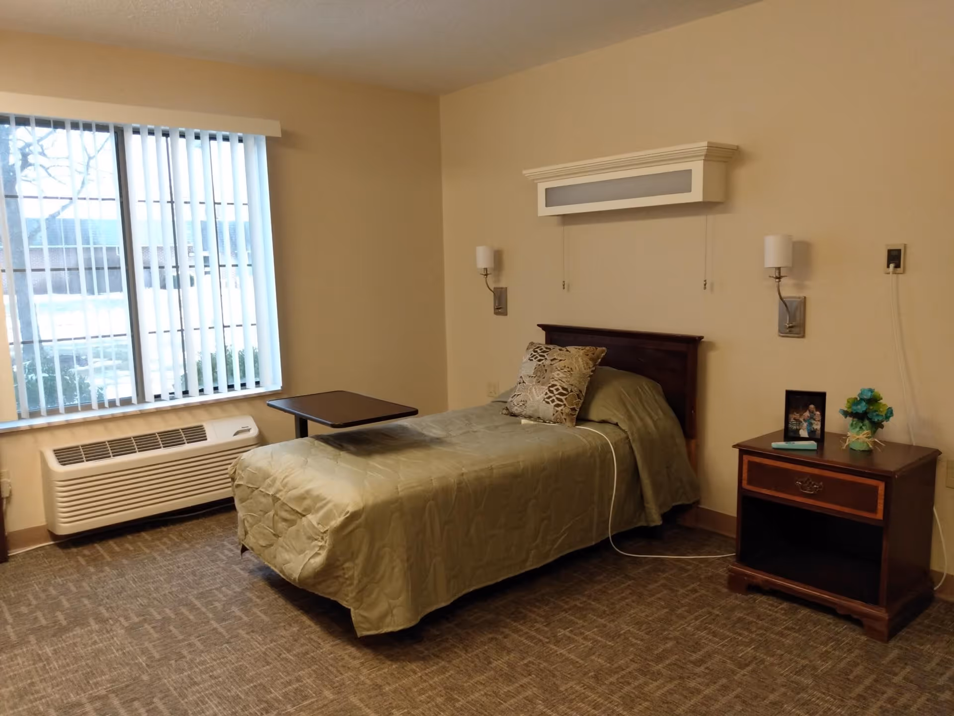 A simple bedroom in an assisted living facility featuring a single bed with a green bedspread and a decorative pillow. Next to the bed is a wooden nightstand with a framed photo and a small vase with blue flowers. The room has beige walls, a window with vertical blinds, a wall-mounted air conditioning unit below the window, and two wall-mounted lamps on either side of the bed.