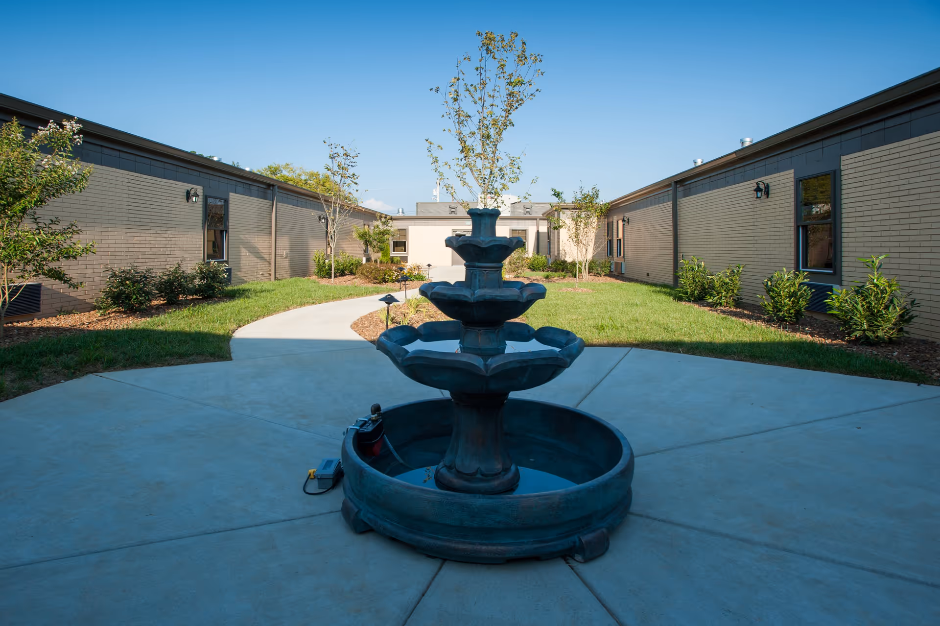Outdoor courtyard area with a three-tiered water fountain in the center, surrounded by a concrete pathway and green grass with small trees and shrubs. The courtyard is enclosed by single-story brick buildings under a clear blue sky.