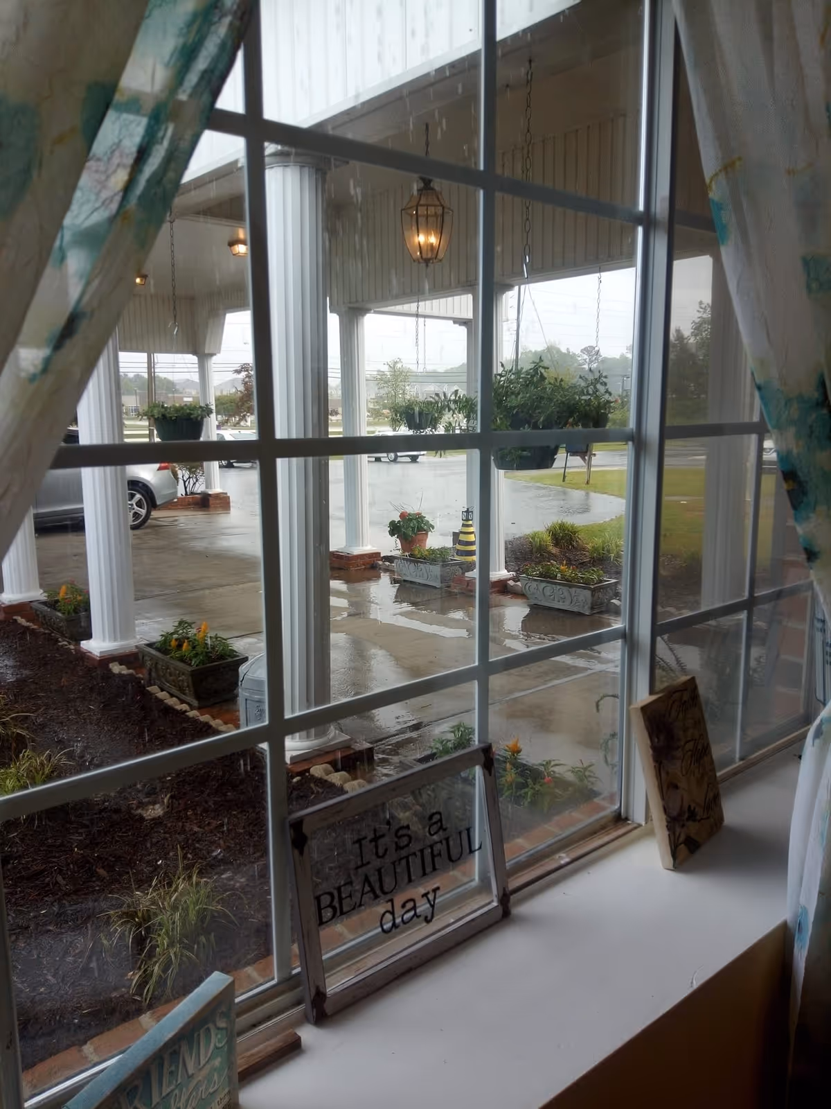View through a window showing a covered entrance with white columns, hanging plants, wet driveway, and a windowsill sign reading "It's a BEAUTIFUL day".