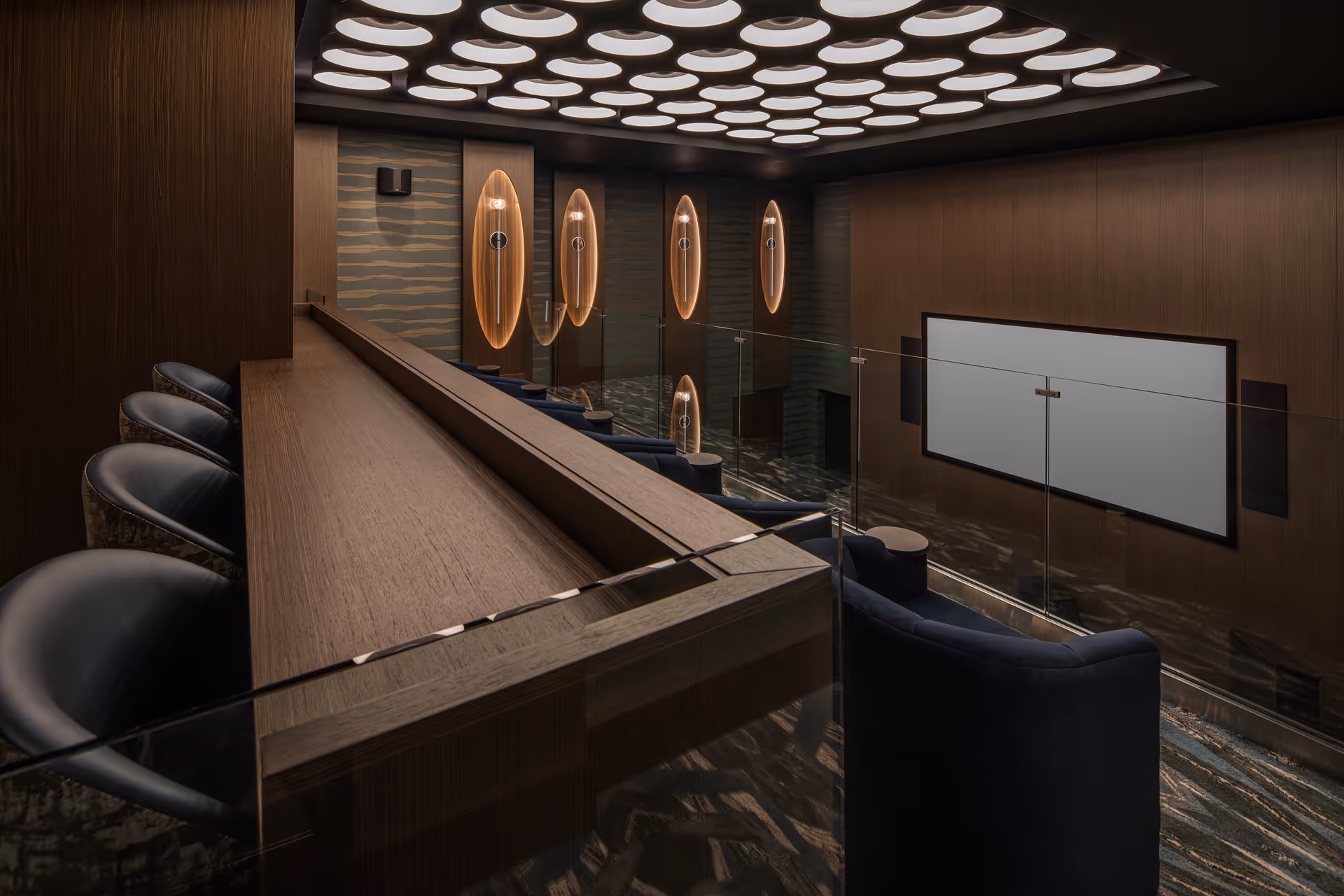 Modern theater room with tiered seating featuring dark blue armchairs and a wooden counter with black leather chairs. The room has wood-paneled walls, a large blank screen, and a ceiling with circular recessed lights. Decorative oval wall lights are mounted on the far wall.