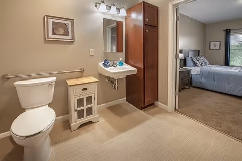 A clean bathroom with a white toilet, a small wooden cabinet, a wall-mounted sink with soap and a towel, a mirror, and a light fixture above. The bathroom has beige walls and flooring. Through an open doorway, a bedroom with a bed, nightstand, lamp, and window with blinds is visible.
