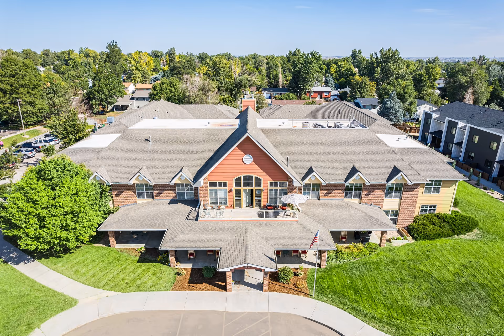 Aerial view of a large assisted living facility building with a gray shingled roof, red and yellow exterior walls, and multiple windows. The building has a covered entrance with an American flag on a flagpole nearby. Surrounding the building are well-maintained green lawns, trees, and a parking area with several cars. The sky is clear and blue.