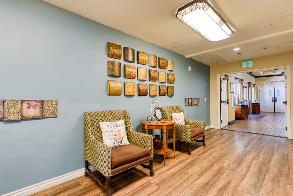 A cozy interior seating area with two patterned armchairs featuring floral pillows, a small wooden side table with a decorative clock, and wall art consisting of multiple square metal pieces arranged in a grid on a blue accent wall. The floor is wood, and there is a hallway leading to double doors labeled 'Legacies'.