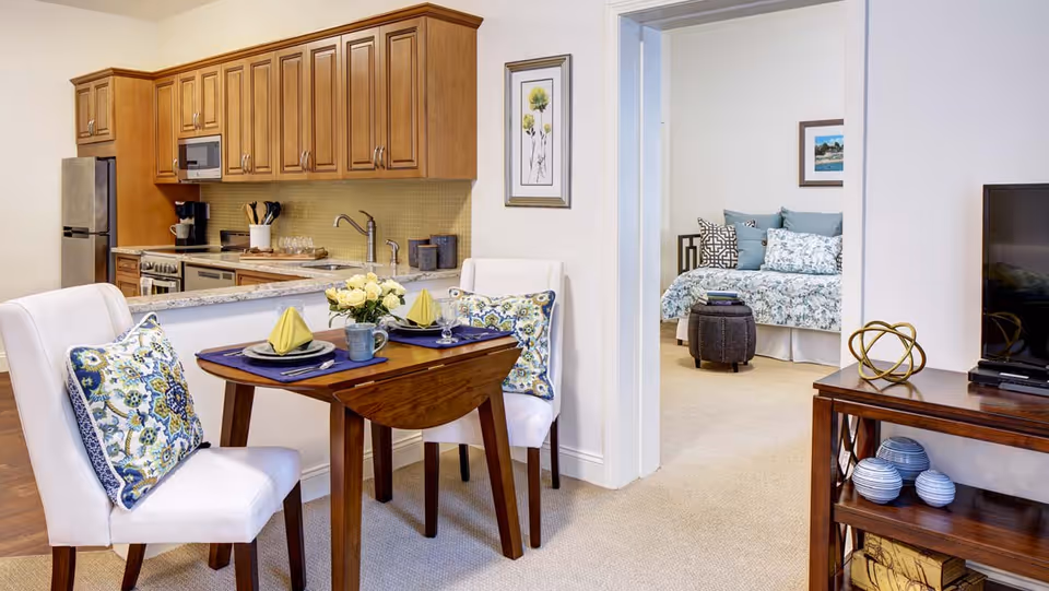 A cozy dining area with a small wooden table set for two, featuring white cushioned chairs with patterned pillows. The dining area is adjacent to a kitchen with wooden cabinets, stainless steel appliances, and a granite countertop. In the background, a bedroom with a floral bedspread and decorative pillows is visible through an open doorway. A wooden TV stand with decorative items and a flat-screen TV is on the right side of the image.