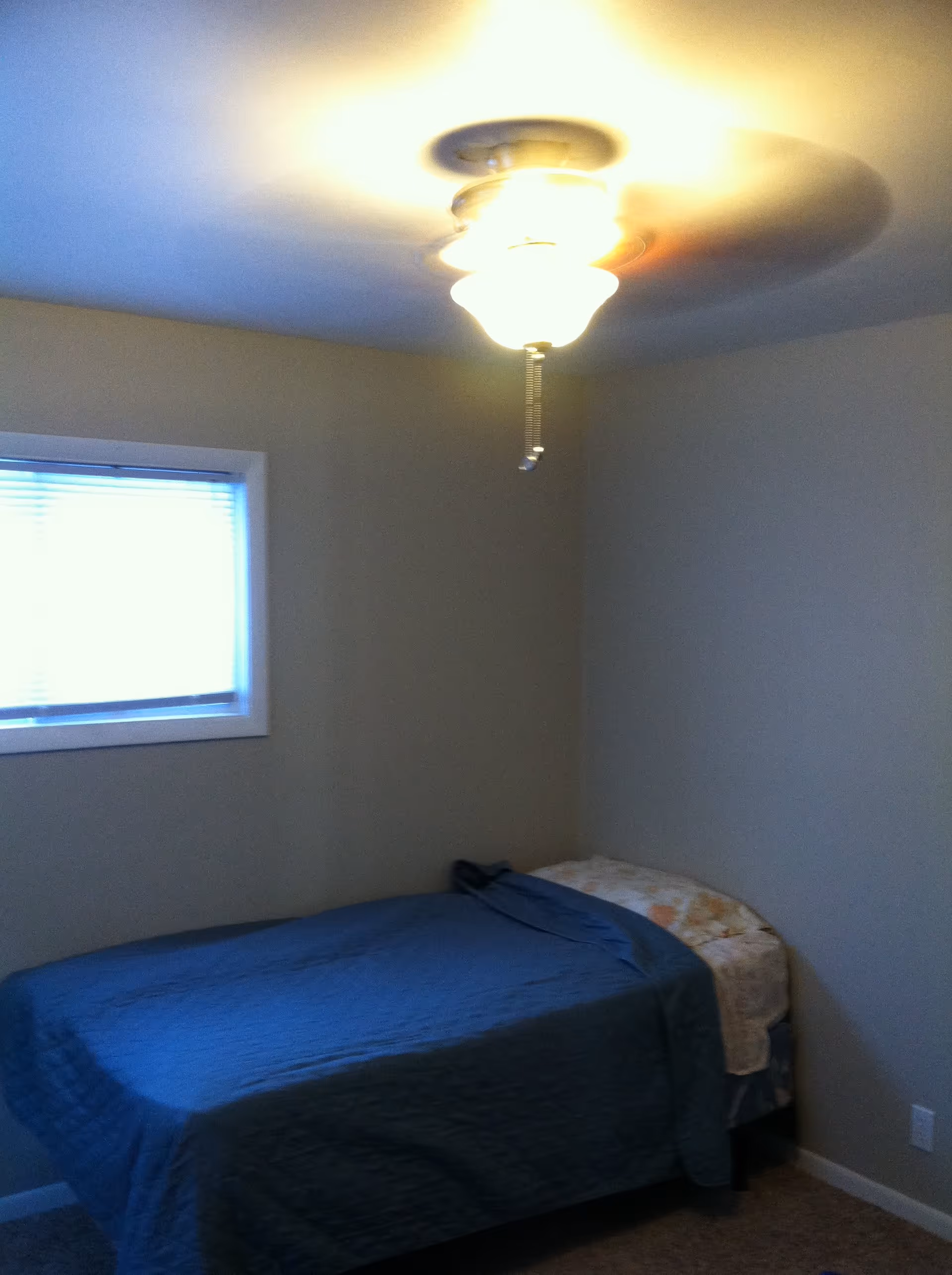 A small bedroom with a single bed covered with a blue blanket and a floral pillow. There is a window with blinds on the left wall and a ceiling light fixture with a pull chain turned on.