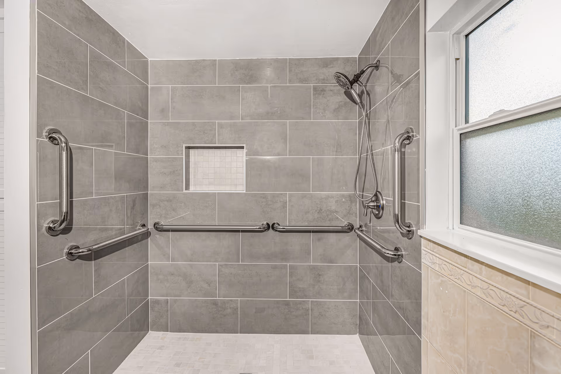 A spacious walk-in shower with gray tiled walls and a white tiled floor. The shower features multiple stainless steel grab bars on three walls for safety and accessibility. There is a built-in recessed shelf in the wall for toiletries and a handheld showerhead mounted on the right wall. A frosted window is visible on the right side, allowing natural light into the shower area.