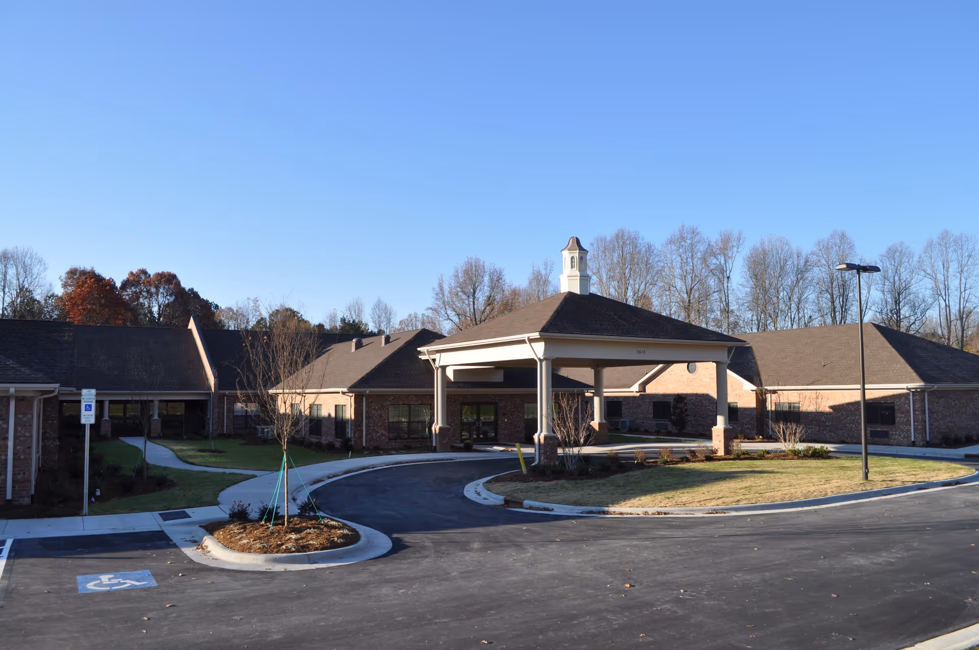 Exterior view of a single-story brick building with a covered entrance and a circular driveway, surrounded by young trees and a parking lot with a handicapped parking space, under a clear blue sky.